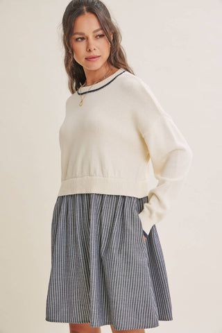 A woman modeling a cream knit sweater with a black and white patterned crew neck, paired with a blue and white vertically striped pleated skirt, against a solid light beige background.