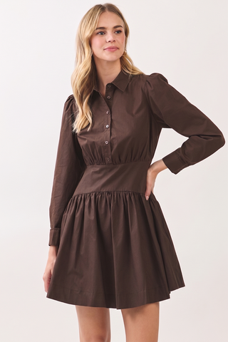 A woman modeling a dark brown long-sleeve mini dress with a button-down collar, gathered waist, and flared skirt against a plain light background.