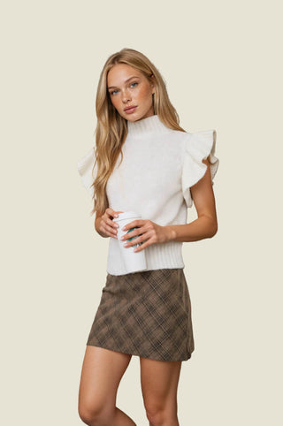 A woman stands indoors against a stone wall wearing a white mock-neck top with ruffled cap sleeves and a brown plaid mini skirt, holding a white coffee cup.