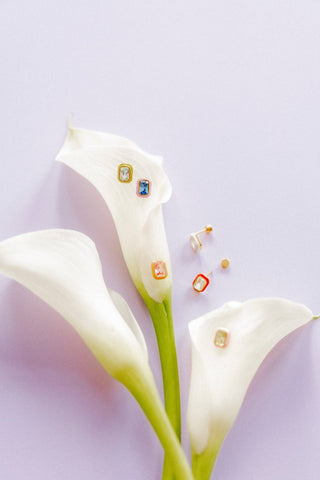 Gold earrings with gemstones on a white flower against a light purple background