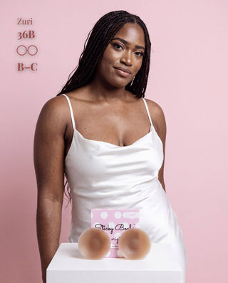 A woman wearing a white camisole stands against a pink background, with two freestanding nude silicone bra cups on a white pedestal in front of her and text in the upper left reading 'Zuri 36B B-C'.