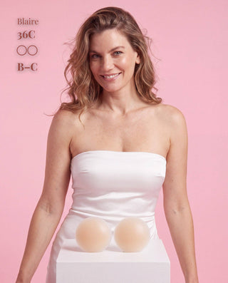A woman stands against a pink studio background wearing a white strapless tube top, with two nude silicone bra cups displayed on a white pedestal in front of her. Text on the upper left reads 'Blaire 36C OO B-C'.