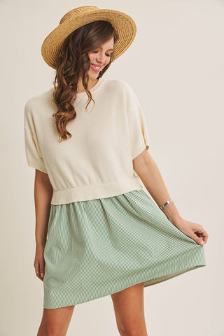 A woman wearing a cream short-sleeve knit top and a light green and white striped mini skirt, paired with a straw hat, posing against a solid light beige background.