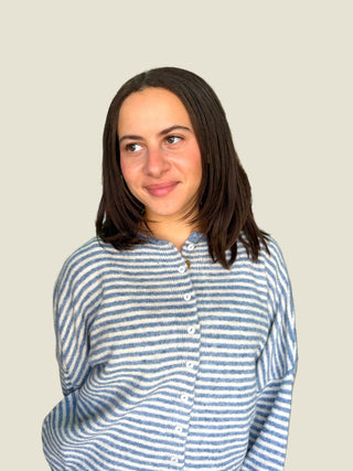 A woman with shoulder-length dark brown hair wearing a blue and white striped button-up henley shirt, photographed against a solid light beige background.