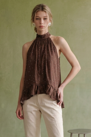 A woman modeling a brown halter-neck top with a small white polka dot pattern and a ruffled hem, paired with cream pants, set against a textured olive green wall.