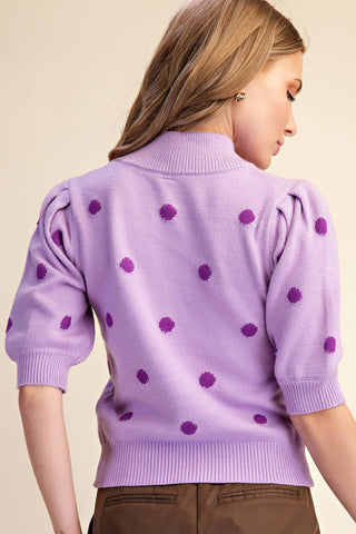 A woman shown from the back wearing a lavender purple knit sweater with darker purple polka dots and puff sleeves, paired with brown pants, against a solid beige background.