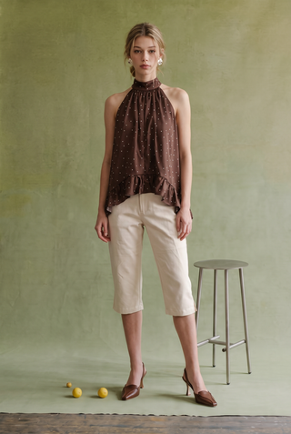 A woman stands in a studio wearing a sleeveless high-neck brown top with small white dots and cream cropped pants, paired with brown pointed-toe heels. A green textured backdrop, a small stool, and a few yellow objects on the floor are visible.