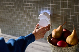 Person holding a Pura smart fragrance diffuser against a tiled wall with a bowl of pears on a table.