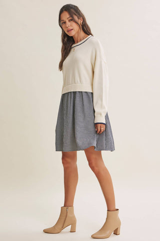 A woman modeling a cream knit sweater and a blue and white striped pleated mini skirt, paired with beige ankle boots, against a solid light beige background.