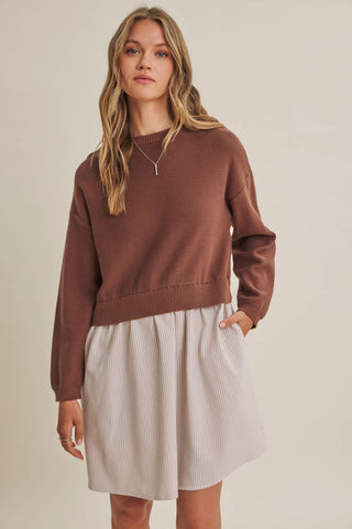 A woman wearing a brown knit crewneck sweater and a light beige pleated skirt, standing against a plain beige studio background.