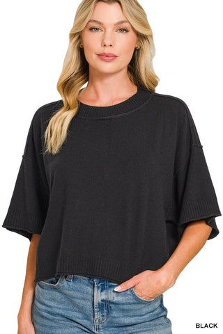 A woman wearing a black short-sleeve knit top with a relaxed, oversized fit and ribbed crew neckline, paired with blue jeans, against a plain white background.