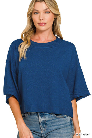 A woman wearing a blue knit short-sleeve top with drop shoulders and ribbed trim, paired with blue jeans, against a plain white background.