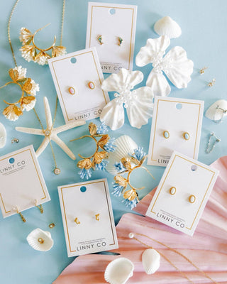 A styled flat-lay of multiple LINNY CO stud earrings displayed on white cards, surrounded by seashells, a starfish, floral decorations, and soft pink fabric on a light blue background.