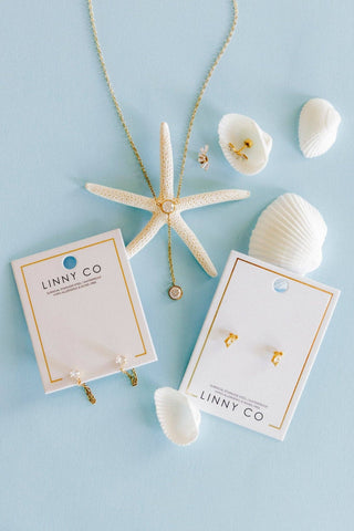 A flat-lay of jewelry from the brand LINNY CO on a light blue background, featuring a gold necklace with a starfish pendant and matching gold stud earrings on white display cards, styled with white seashells.