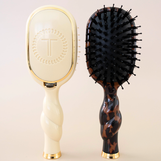 Two paddle hairbrushes shown side by side on a light beige studio background. The left brush has an oval cream face with gold trim and a twisted handle with gold accents.