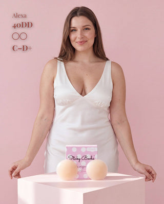 A woman in a white satin slip dress stands behind a pink pedestal displaying two beige sticky boobs and a pink box labeled 'Sticky Boobs'. Text on the top left reads 'Alexa 40DD C-D+'.