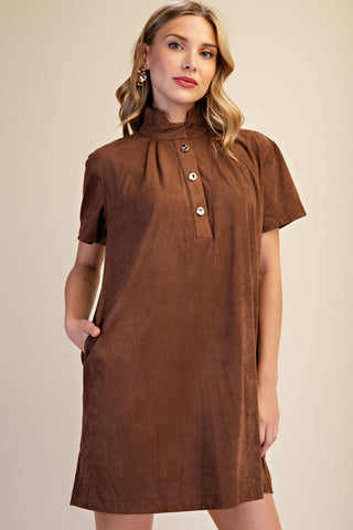 A woman modeling a brown short-sleeve shirt dress with a high gathered neckline and three decorative buttons, set against a plain beige studio background.
