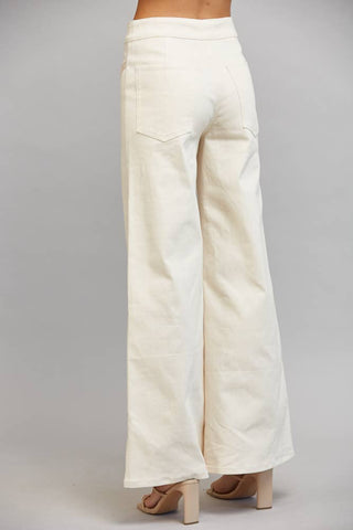 A person shown from the back wearing off-white wide-leg pants and beige block-heel sandals against a plain light gray studio background.