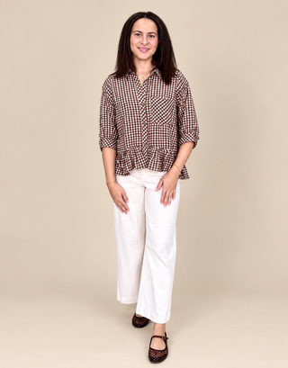 A woman modeling a brown and white checkered button-down shirt with a ruffled hem, paired with white wide-leg pants and dark brown shoes, against a solid beige background.