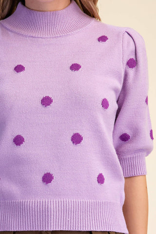 A close-up of a lavender knit sweater with darker purple textured polka dots, featuring a mock neck and short puff sleeves, shown on a model against a light beige background.