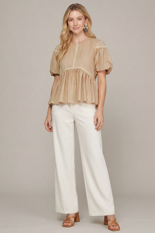 A woman modeling a beige short-sleeve peplum blouse with puff sleeves and white wide-leg pants, standing against a neutral light gray studio background.
