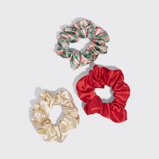 Three fabric hair scrunchies on a light gray background: one cream satin scrunchie, one red satin scrunchie, and one green and red plaid scrunchie.
