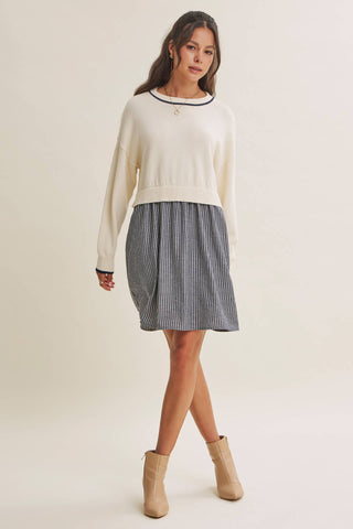 A woman modeling a cream knit sweater with a black crew neck trim, paired with a gray and white vertically striped pleated skirt and beige ankle boots, against a solid light beige studio background.