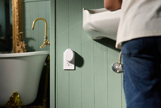 A white circular smart plug device shown mounted on a sage green wood-paneled bathroom wall next to a sink and faucet, with part of a person in a white shirt and blue jeans visible on the right.
