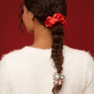 A woman shown from the back wearing a white fuzzy sweater, with her dark brown hair styled in a braid. She is wearing two fabric scrunchies: a bright red one near the crown and a green, white, and red plaid one at the end of the braid, set against a solid dark red background.