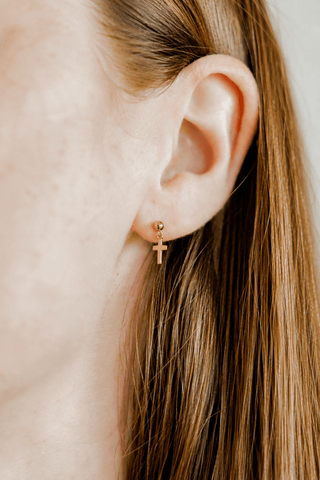A close-up of a person's ear wearing a small gold cross stud earring, with light brown hair visible against a soft light gray background.