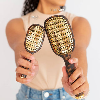 Person holding a mini and full-size hairbrush with gold bristle heads against a neutral background.