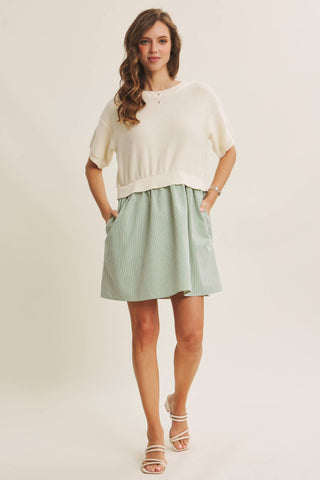 A woman modeling a cream short-sleeve knit top and a light sage green mini skirt with side pockets, paired with white strappy sandals, against a plain cream studio background.