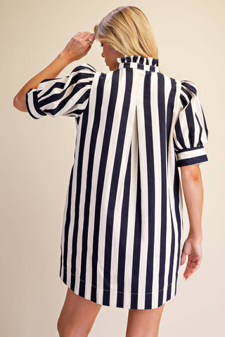 A woman shown from the back wearing a short puff-sleeve shirt dress with bold vertical navy and white stripes, set against a plain beige studio background.