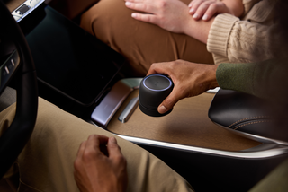 A close-up lifestyle shot of a person holding a black cylindrical smart speaker with a glowing blue ring in a car's center console next to a tablet and phone.