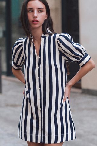 A woman wearing a navy and off-white vertically striped puff-sleeve shirt dress, standing outdoors with her hands on her hips.