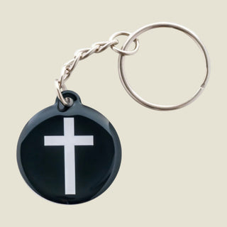 A round black glossy keychain featuring a white Christian cross in the center, attached to a silver metal chain and split key ring, shown on a plain white background.