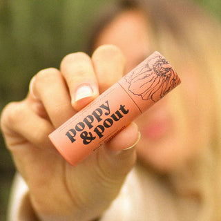 Hand holding a pink lip balm with 'poppy & pout' branding against a blurred background