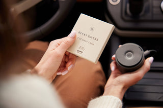 Person holding a BECKI OWENS product and a black container inside a vehicle.