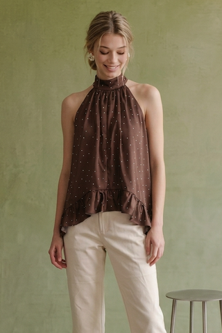 A woman wearing a brown halter-neck top with a small white polka dot pattern and cream pants, standing against a textured sage green wall.