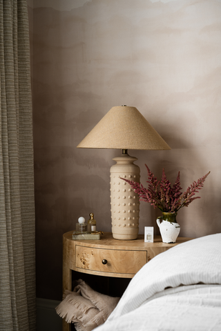 A bedside table in a softly styled bedroom setting, featuring a textured cream ceramic table lamp with a beige conical shade, a small white pitcher with dried burgundy flowers, and other decorative accents against a warm, textured wall. Studio Megee Sea Salt Driftwood fragrancei refill bottle and packaging on a beige background