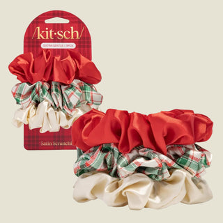 A set of four Kit Sch satin scrunchies displayed on a light gray background, with the retail packaging card standing behind them. The scrunchies include a large red satin scrunchie, a green and red plaid satin scrunchie, a cream satin scrunchie, and a matching plaid and satin combination.