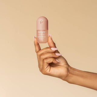 A hand with pale pink manicured nails holds a small bottle of Kitsch Cosmetics liquid shimmer against a solid beige background.