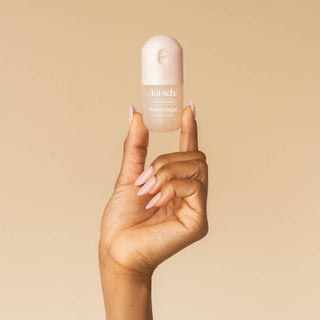 A hand with pale pink manicured nails holds a small Kit-schi Warm Sugar bottle against a solid warm beige background.