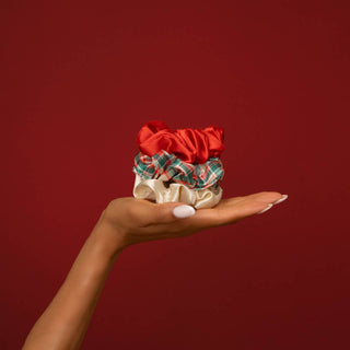 Three fabric hair scrunchies in red, green plaid, and cream are stacked on an open hand against a solid dark red background.