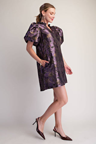A woman models a short, puffed-sleeve mini dress with a purple and gold floral jacquard pattern, paired with dark brown pointed-toe slingback heels against a light gray studio background.