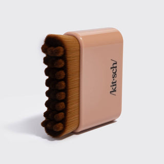 A compact makeup brush with a peach-pink handle and brown bristles, featuring the black printed brand name 'Kit-sch' on the side, set against a light gray background.