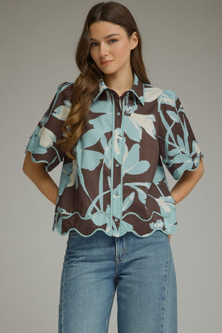 A woman modeling a short-sleeve button-up blouse with a brown, light blue, and white floral pattern, paired with blue jeans, against a solid gray studio background.