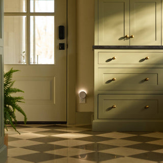pura plugged into a wall by Green kitchen cabinets with gold handles in a room with a checkered floor and a plant.