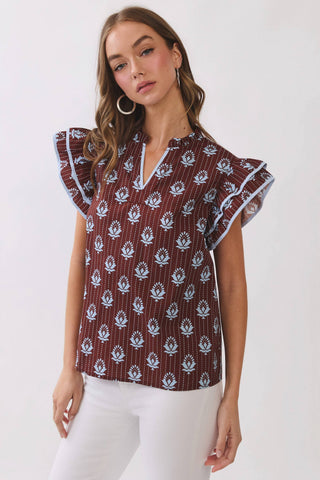 A woman modeling a short-sleeve blouse with flutter sleeves, featuring a brown base with white floral block prints and thin vertical stripes, paired with white pants against a plain white studio background.