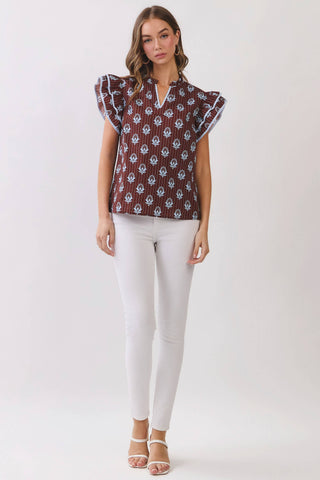 A woman modeling a short-sleeve top with flutter sleeves, featuring a brown base with a white floral ikat pattern, paired with white pants and beige sandals, set against a plain light gray studio background.
